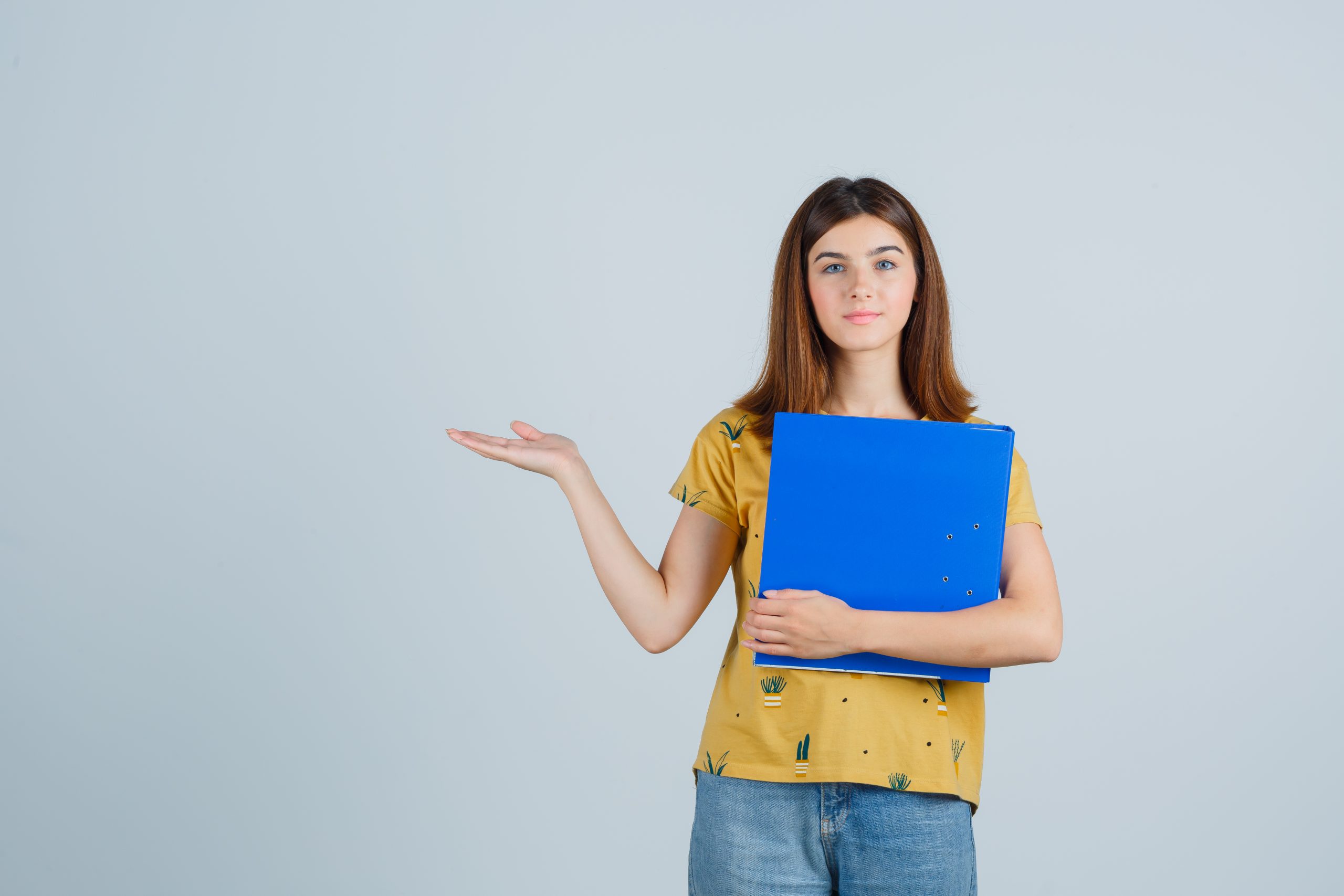 young girl holding file folder, spreading palm aside in yellow t-shirt and jeans and looking buoyant. front view.